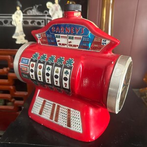May include: A red Barney's slot machine bank with a black cap on top. The front features five spinning reels displaying shamrocks, sevens, and fruit symbols. A payout chart is printed on the upper section, and a coin slot chart is on the base.