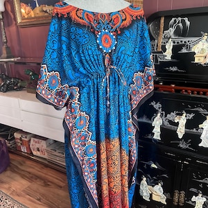 May include: A vibrant blue, orange, and black patterned caftan dress. The dress features a round neckline, a drawstring waist, and wide sleeves. The fabric is adorned with intricate paisley and floral designs, creating a bohemian aesthetic. The dress is displayed on a mannequin.