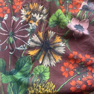 May include: A floral pattern on a maroon fabric. The design features detailed illustrations of various flowers in shades of orange, yellow, pink, and white, with green leaves and stems. The fabric has a slightly wrinkled texture.
