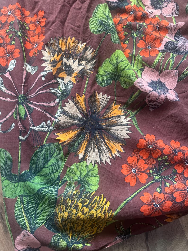 May include: A floral pattern on a maroon fabric. The design features detailed illustrations of various flowers in shades of orange, yellow, pink, and white, with green leaves and stems. The fabric has a slightly wrinkled texture.
