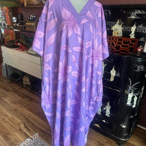 May include: A floor-length, loose-fitting kaftan dress in a vibrant lavender shade, featuring a repeating pattern of large, stylized flowers and leaves in a slightly darker purple tone. The garment has a V-neckline and short, wide sleeves.
