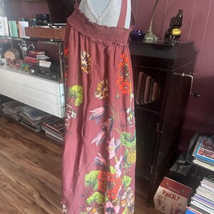 May include: A maroon maxi dress with a floral pattern. The dress features a smocked bodice and wide straps. The floral design includes red, orange, yellow, and green flowers and leaves. The dress is displayed on a mannequin.