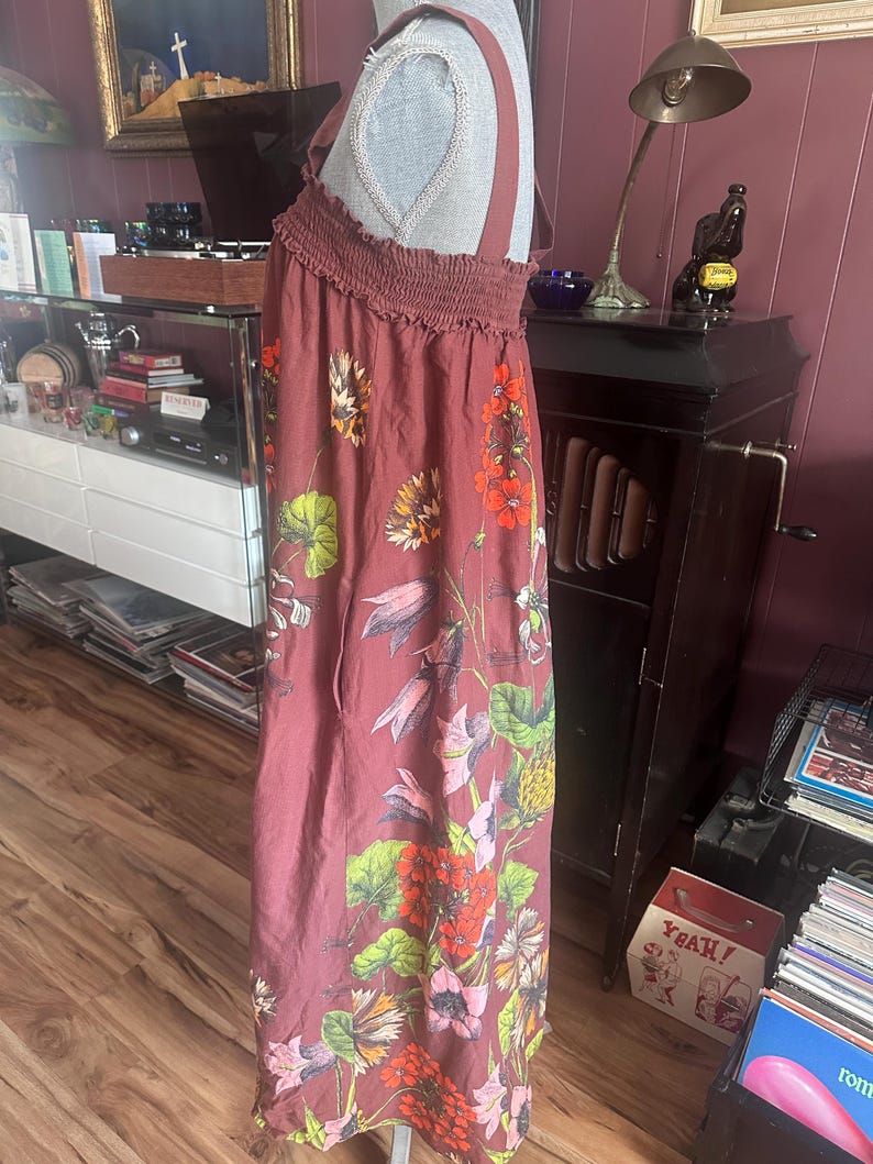 May include: A maroon maxi dress with a floral pattern. The dress features a smocked bodice and wide straps. The floral design includes red, orange, yellow, and green flowers and leaves. The dress is displayed on a mannequin.