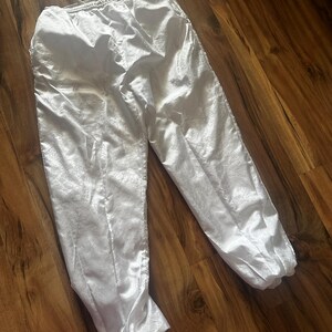 Op de afbeelding: Witte sportbroek met een elastische tailleband en trekkoord. De broek heeft een glanzende, zijdeachtige textuur en is bij de enkels gerimpeld. De broek ligt op een houten oppervlak.