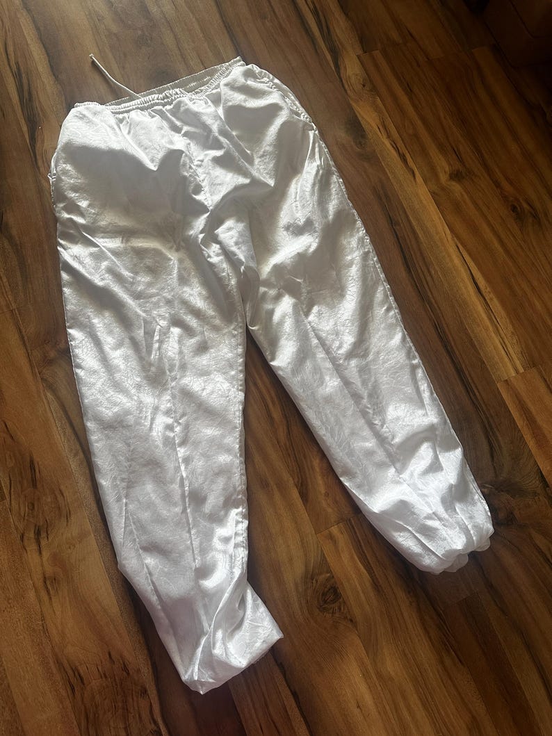 Op de afbeelding: Witte sportbroek met een elastische tailleband en trekkoord. De broek heeft een glanzende, zijdeachtige textuur en is bij de enkels gerimpeld. De broek ligt op een houten oppervlak.