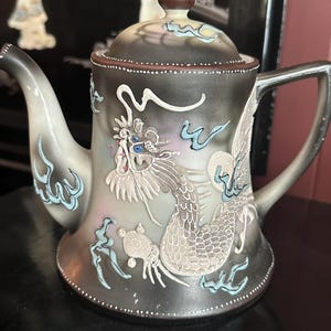 May include: A ceramic teapot featuring a gradient black and grey finish with a raised, white relief dragon design. The dragon has blue eyes and is surrounded by light blue cloud motifs. The teapot includes a matching lid with a brown knob.