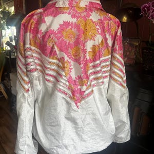 Op de afbeelding: Een witte jas met een bloemenpatroon van roze en gele bloemen op de bovenrug en schouders. De jas heeft een kraag en lange mouwen. Het onderste deel van de jas is wit.