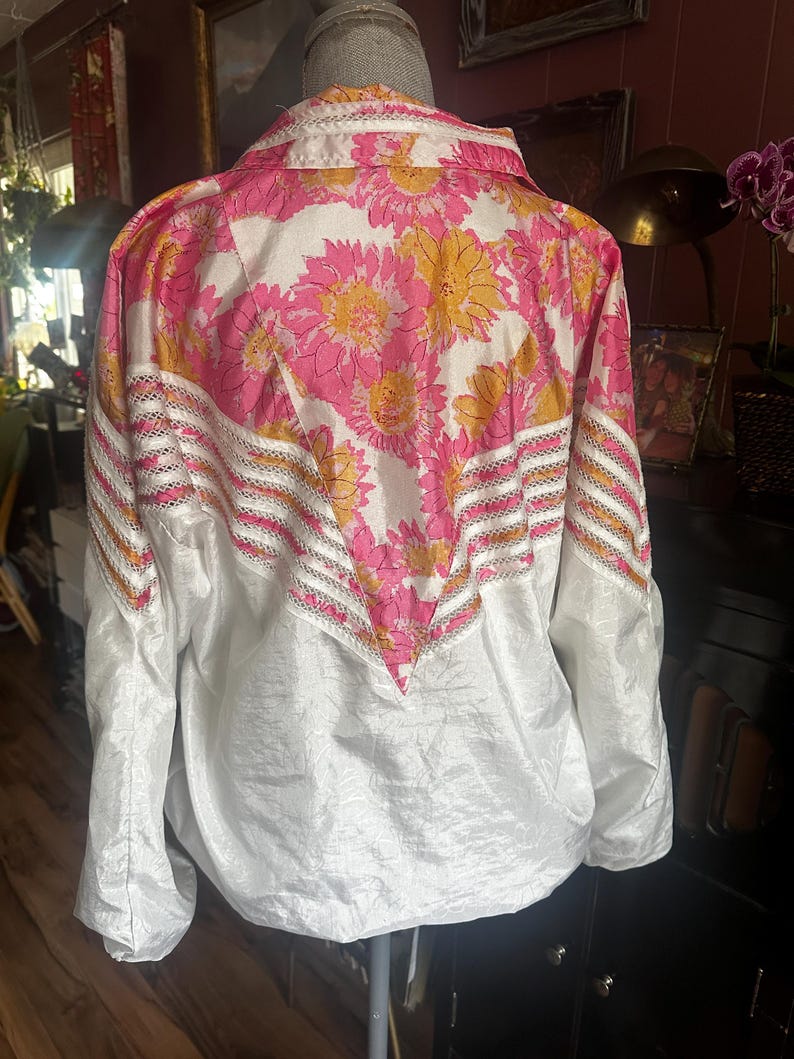 Op de afbeelding: Een witte jas met een bloemenpatroon van roze en gele bloemen op de bovenrug en schouders. De jas heeft een kraag en lange mouwen. Het onderste deel van de jas is wit.