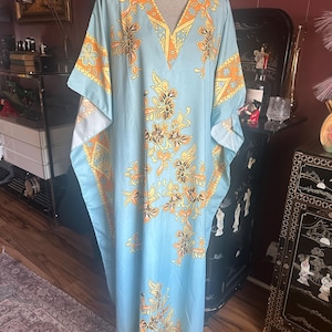May include: A floor-length light blue kaftan dress featuring a vibrant orange and yellow floral pattern. The garment has a deep V-neckline with a wide orange border and wide, flowing sleeves that extend from the shoulders to the hemline.