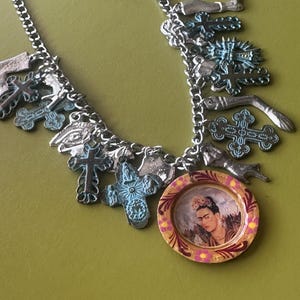Könnte beinhalten: Silberfarbene Halskette mit verschiedenen Anhängern, darunter Kreuze, Fische und ein runder Anhänger mit einem Porträt von Frida Kahlo. Der Anhänger hat einen floralen Rand. Die Anhänger haben ein Vintage-Aussehen.