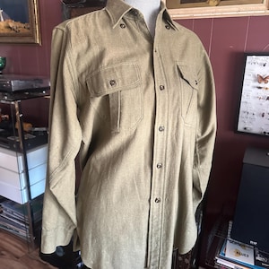 May include: A long-sleeved button-down shirt in a muted olive green color. The shirt features a standard collar, two chest pockets with buttoned flaps, and a front placket with matching buttons. The fabric has a visible, slightly textured weave.