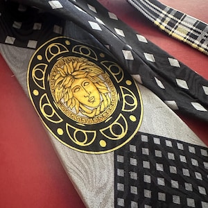 May include: A black and gray silk tie featuring a gold Medusa head emblem. The tie has a geometric pattern with diamond shapes and a plaid design on the narrow end. The tie is a classic accessory.