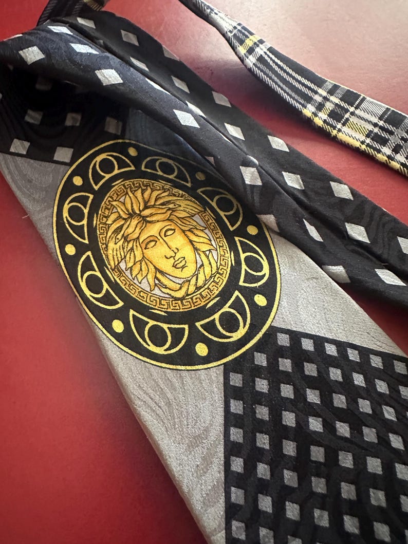 May include: A black and gray silk tie featuring a gold Medusa head emblem. The tie has a geometric pattern with diamond shapes and a plaid design on the narrow end. The tie is a classic accessory.