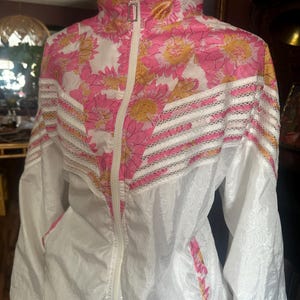 Op de afbeelding: Witte jas met roze en roze bloemenprint, hoge kraag en volledige ritssluiting. De jas heeft een roze en gele bloemenpatroon op de bovenborst en kraag, met witte en kanten accenten. De jas heeft twee zakken en een trekkoord in de zoom.