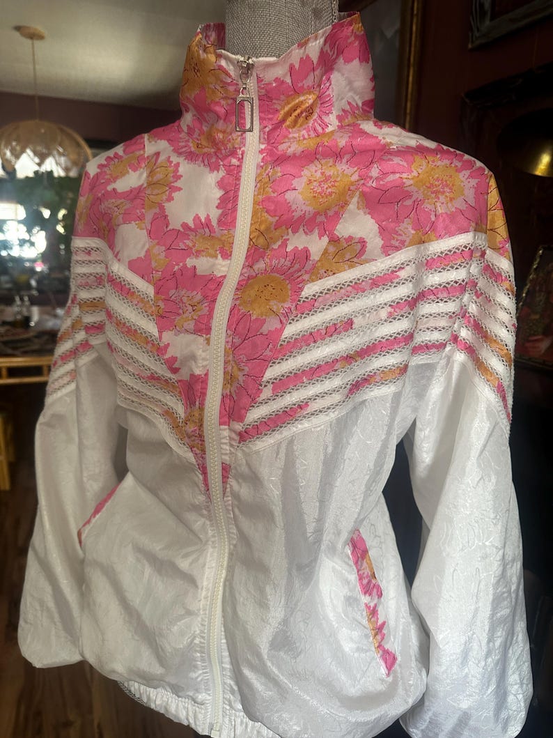 Op de afbeelding: Witte jas met roze en roze bloemenprint, hoge kraag en volledige ritssluiting. De jas heeft een roze en gele bloemenpatroon op de bovenborst en kraag, met witte en kanten accenten. De jas heeft twee zakken en een trekkoord in de zoom.