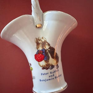 May include: White ceramic basket with a handle, featuring a Peter Rabbit and Benjamin Bunny illustration. The basket has gold trim and the text "Peter Rabbit and Benjamin Bunny" on the side. The background is a solid red.