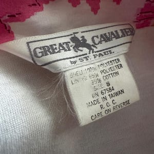 Op de afbeelding: Close-up van een kledinglabel. Het label is wit met zwarte tekst en een logo van een paard en ruiter. Het label leest "GREAT CAVALIER by ST. PAUL" en vermeldt de materialen als polyester en katoen. Het label zegt ook "MADE IN TAIWAN".
