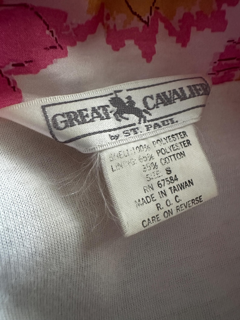 Op de afbeelding: Close-up van een kledinglabel. Het label is wit met zwarte tekst en een logo van een paard en ruiter. Het label leest "GREAT CAVALIER by ST. PAUL" en vermeldt de materialen als polyester en katoen. Het label zegt ook "MADE IN TAIWAN".