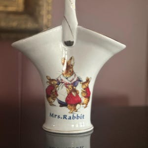 May include: White ceramic basket with a handle, featuring a Mrs. Rabbit illustration with her children in red coats. The basket has a gold trim and the words "Mrs. Rabbit" are printed on the front.