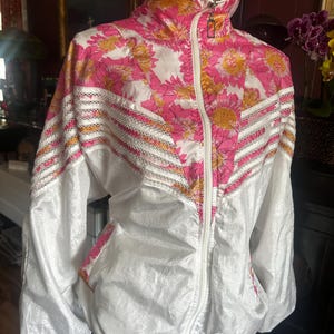 Op de afbeelding: Een vintage witte en roze jas met bloemenprint, een hoge kraag en een ritssluiting over de hele lengte. De jas heeft een levendig bloemenpatroon in roze en geel, met witte accenten en een decoratieve rand.
