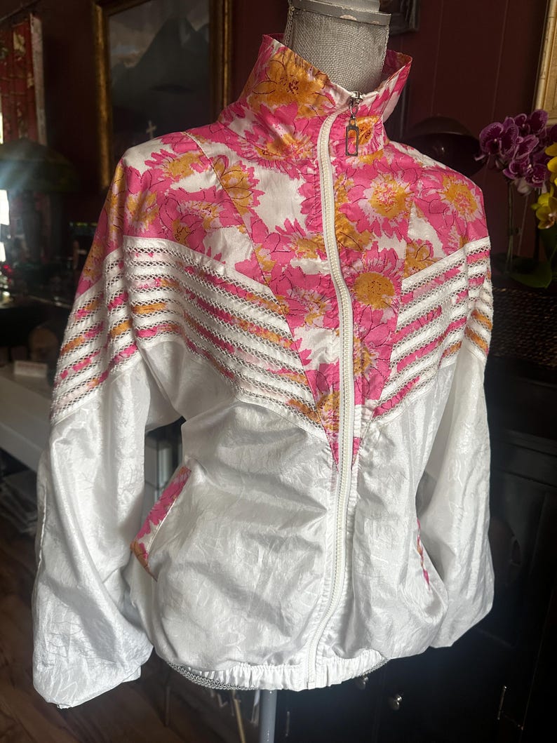 Op de afbeelding: Een vintage witte en roze jas met bloemenprint, een hoge kraag en een ritssluiting over de hele lengte. De jas heeft een levendig bloemenpatroon in roze en geel, met witte accenten en een decoratieve rand.