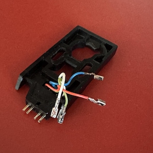 Può includere: Un portatestina per giradischi in plastica nera dotato di quattro piccoli pin metallici a un'estremità e quattro fili sottili codificati a colori con connettori metallici. I fili rosso, blu, bianco e verde si estendono dal telaio.