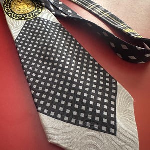 May include: A black and silver tie with a geometric pattern and a gold medallion featuring a face. The tie has a black and white plaid design on the back. The tie is on a red surface.