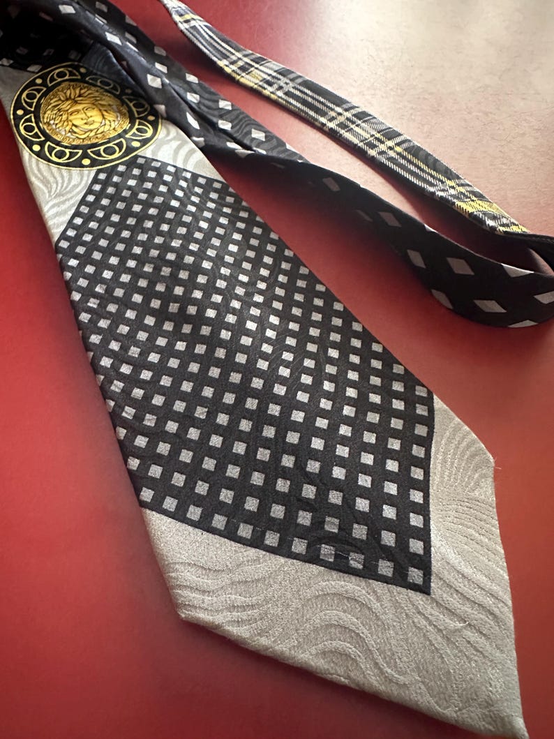 May include: A black and silver tie with a geometric pattern and a gold medallion featuring a face. The tie has a black and white plaid design on the back. The tie is on a red surface.