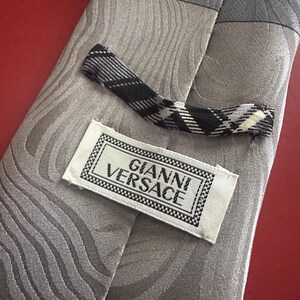 May include: A gray silk tie with a wave pattern and a black and white plaid tip. The label reads "GIANNI VERSACE" in a black and white frame. The tie is on a red surface.