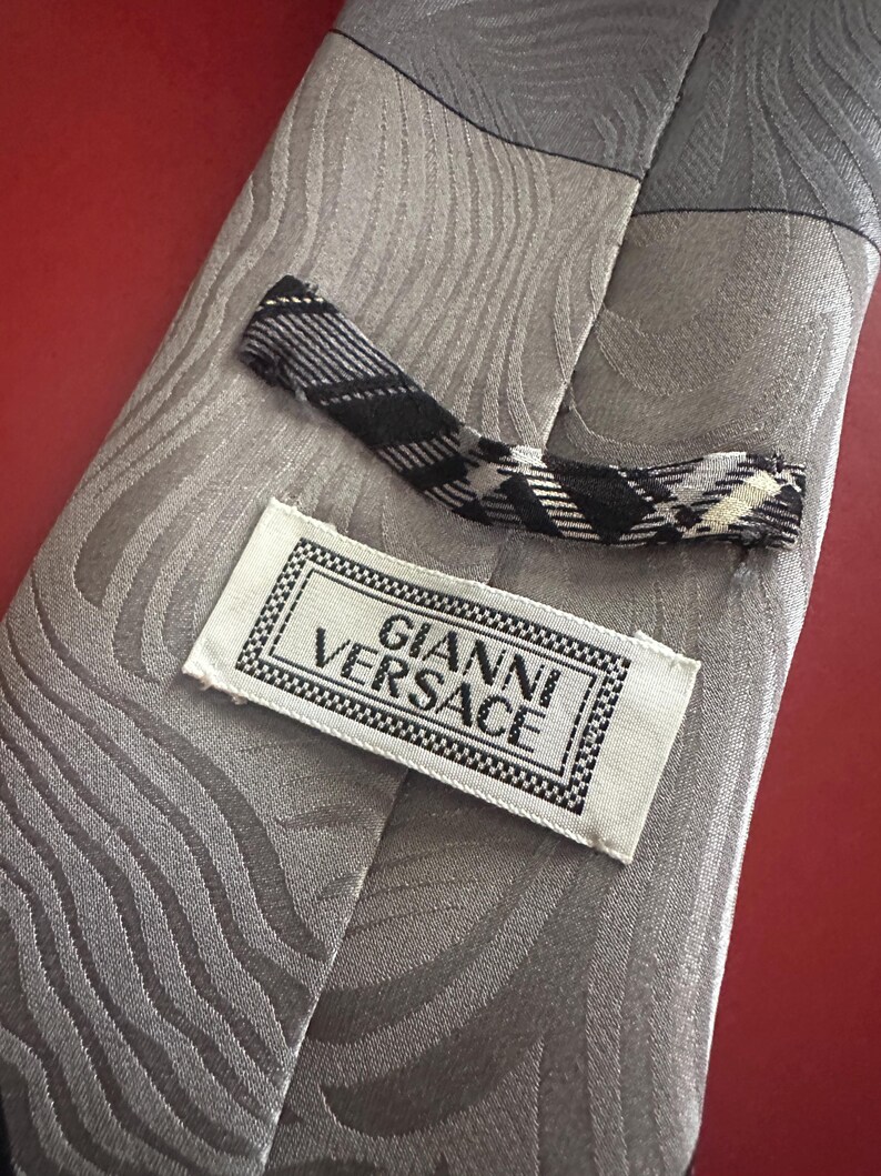 May include: A gray silk tie with a wave pattern and a black and white plaid tip. The label reads "GIANNI VERSACE" in a black and white frame. The tie is on a red surface.