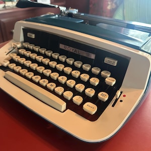 May include: A Royal brand typewriter featuring a cream-colored frame with a dark blue top cover. The device includes a QWERTY keyboard with round cream keys, black letter labels, and specific function keys labeled Magic Column Set and Mar Rel.