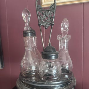 May include: An antique silver-plated condiment set with three clear glass bottles and a small jar. The bottles have decorative stoppers and are held in a silver metal stand with a handle. The base is ornate.