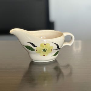 May include: Cream-coloured ceramic gravy boat with a yellow floral design. The gravy boat has a spout, a handle, and is decorated with black and green accents. The floral design is centered on the front of the boat.