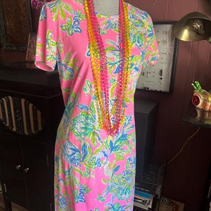 Pode incluir: Um vestido rosa de manga curta com um padr&atilde;o floral em azul, verde e branco. Um colar de contas multicolorido pende em volta do pesco&ccedil;o do manequim. O vestido &eacute; exibido em um manequim.