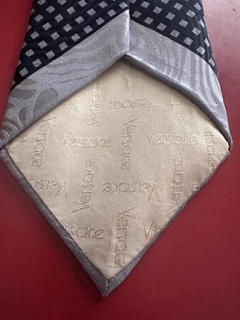 May include: A black and gray patterned necktie with a light beige lining. The lining features the embossed word "Versace" repeated multiple times. The tie's design includes a geometric pattern and a textured border.
