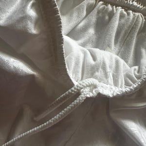 Op de afbeelding: Close-up van een witte sportbroek met een trekkoord. De shorts hebben een zachte, licht glanzende textuur en een comfortabele elastische tailleband. Het trekkoord is wit en gevlochten.