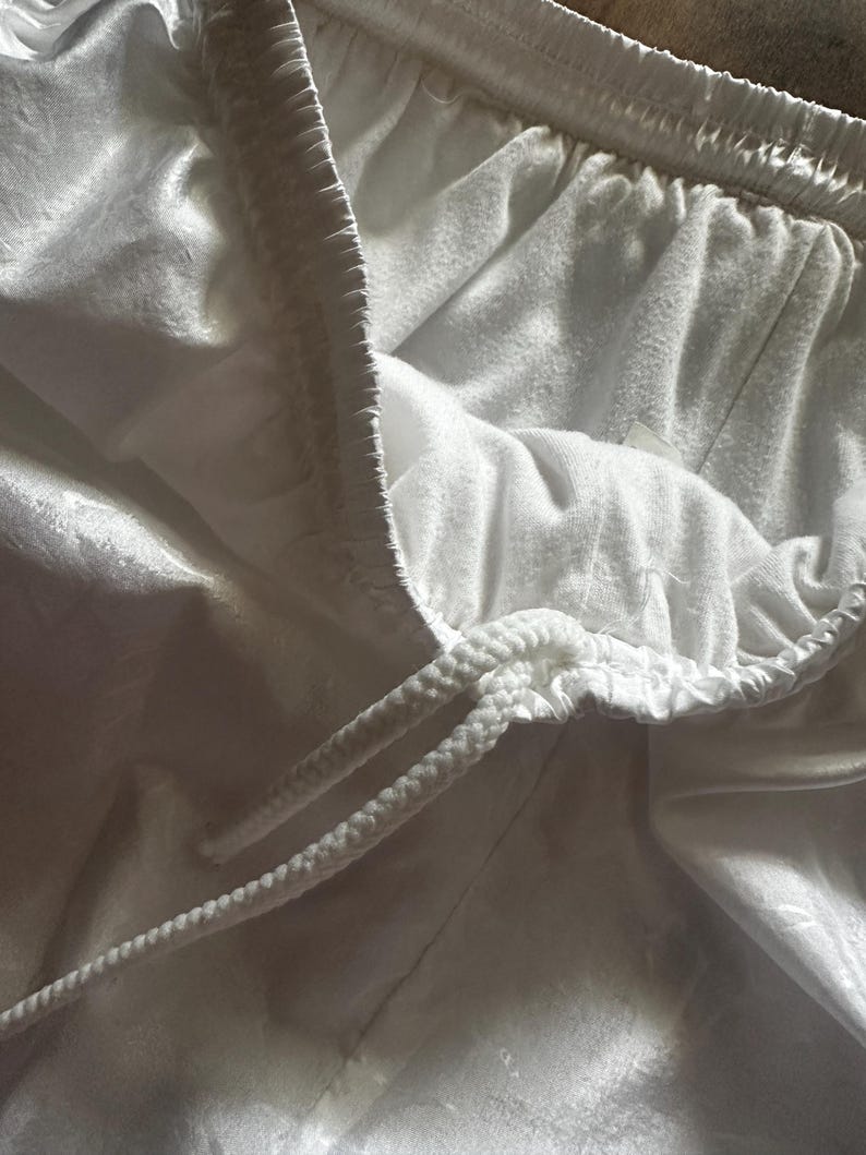 Op de afbeelding: Close-up van een witte sportbroek met een trekkoord. De shorts hebben een zachte, licht glanzende textuur en een comfortabele elastische tailleband. Het trekkoord is wit en gevlochten.