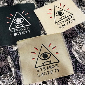May include: Three fabric patches with a black and white illustration of an eye inside a triangle with red rays emanating from the top. The text "STRANGE SOCIETY" is below the triangle.