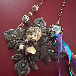 Könnte beinhalten: Eine einzigartige Halskette mit einem detaillierten Cherub-Anhänger mit einem bronzefarbenen Blumendesign. Die Halskette hat eine Kette mit dekorativen Perlen und einen kleineren Anhänger mit bunten Bändern. Strasssteine ​​sorgen für Glanz.
