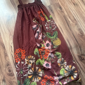 May include: A maroon-colored sundress with floral patterns in shades of orange, pink, and yellow. The dress features thin straps and a gathered neckline. The floral design includes various flower types and green leaves.