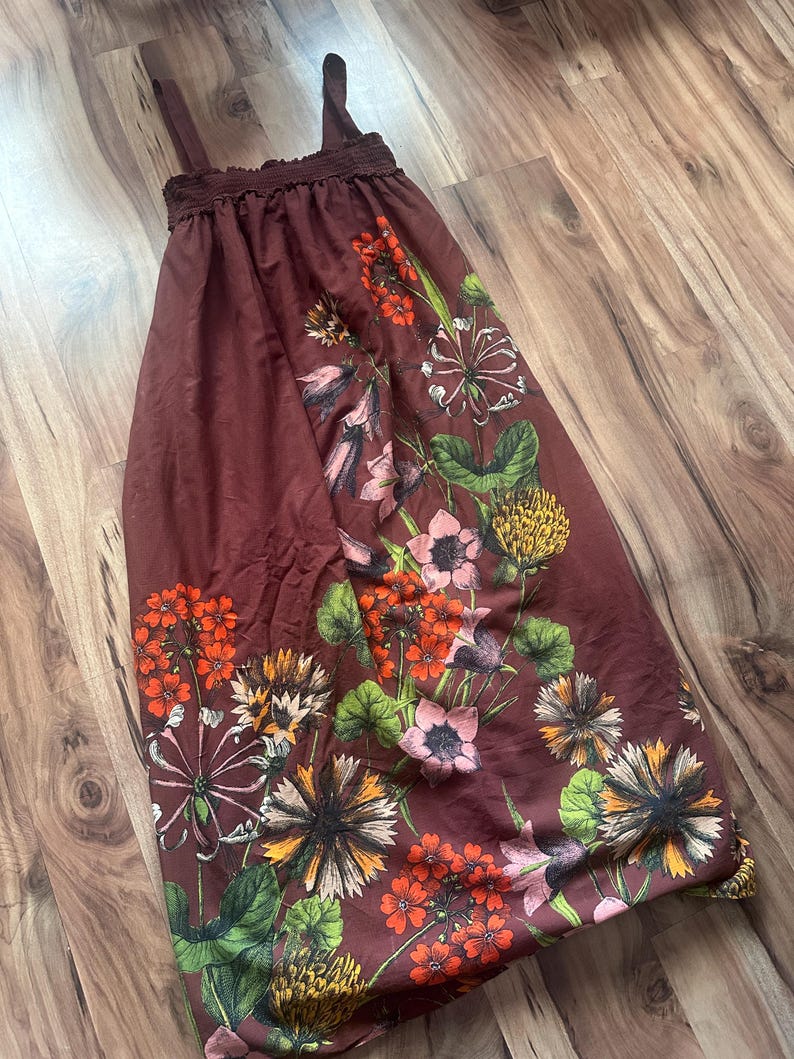 May include: A maroon-colored sundress with floral patterns in shades of orange, pink, and yellow. The dress features thin straps and a gathered neckline. The floral design includes various flower types and green leaves.