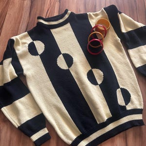 May include: A vintage sweater with a cream and black striped pattern, featuring large half-circle accents. The sweater has a ribbed collar, cuffs, and hem. Several colorful bangles rest on the sweater. The sweater is made of a knit material.