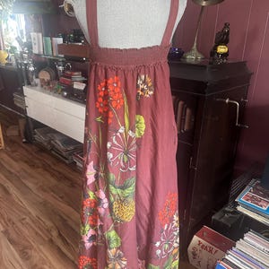 May include: A dusty rose-colored sundress with a floral print. The dress features a smocked bodice and wide shoulder straps. The floral pattern includes orange, pink, and yellow flowers with green leaves. The dress is displayed on a mannequin.