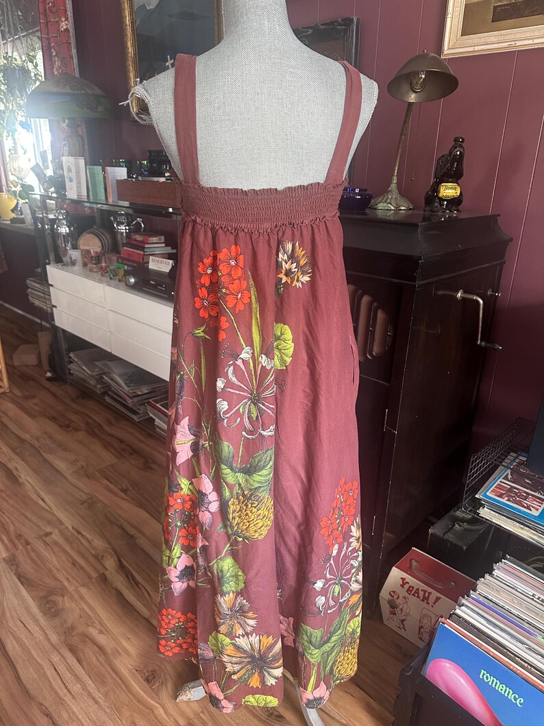 May include: A dusty rose-colored sundress with a floral print. The dress features a smocked bodice and wide shoulder straps. The floral pattern includes orange, pink, and yellow flowers with green leaves. The dress is displayed on a mannequin.