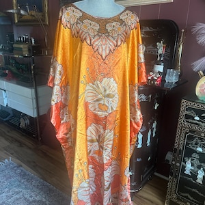 May include: A floor-length kaftan dress in a vibrant orange satin fabric featuring a large-scale floral print in shades of cream and brown. The garment has a wide, loose silhouette with short sleeves and a decorative, intricate brown floral pattern around the neckline.