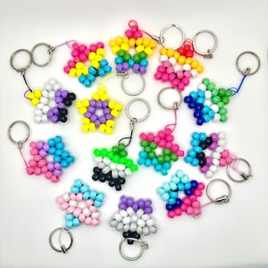 May include: A collection of star-shaped keychains made of colorful beads. Each keychain features a different color scheme, including rainbow, pink, blue, green, and purple. The keychains have silver key rings attached.