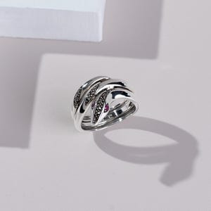 May include: A silver ring with a woven design. The ring features black and red gemstones set into the interwoven bands. The ring is displayed on a white surface with a shadow.