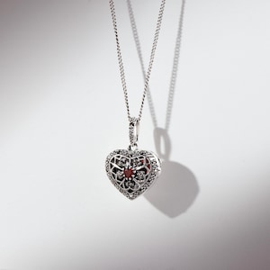 May include: A silver necklace featuring a heart-shaped locket pendant. The locket is decorated with delicate filigree work and a small red gemstone at its centre.