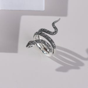 May include: A silver snake ring with a coiled design. The ring features a detailed snake head and body, decorated with small, dark gemstones. The snake's form wraps around the finger, creating a unique and eye-catching piece of jewellery.