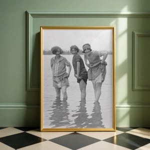 Könnte beinhalten: Gerahmtes Schwarz-Weiß-Foto mit drei Frauen in Vintage-Badebekleidung, die im Wasser stehen und lächeln. Das Foto befindet sich in einem goldenen Rahmen und wird vor einer grünen Wand präsentiert. Ein nostalgisches Bild.